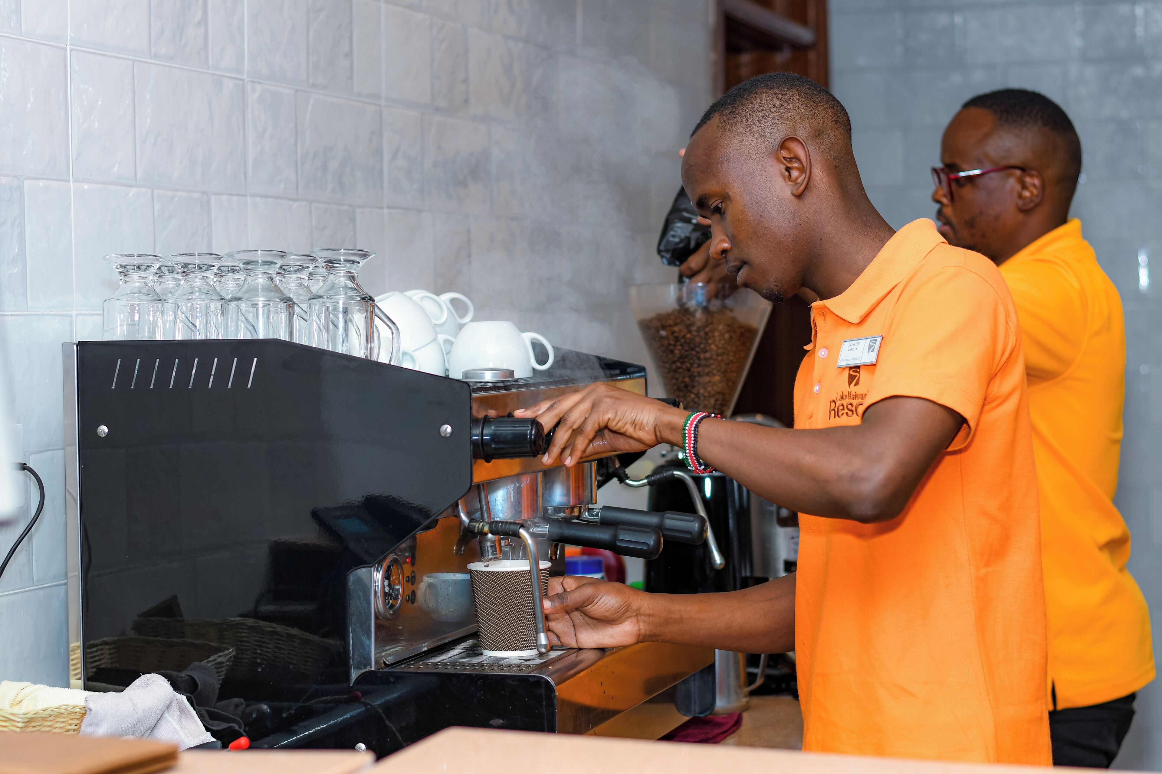 A person operating a coffee machine in a kitchen setting with tiled walls and various kitchen items.