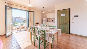 Comedor con una mesa preparada para cuatro, cocina al fondo y una puerta corrediza de vidrio que da a un balcón con mobiliario exterior.