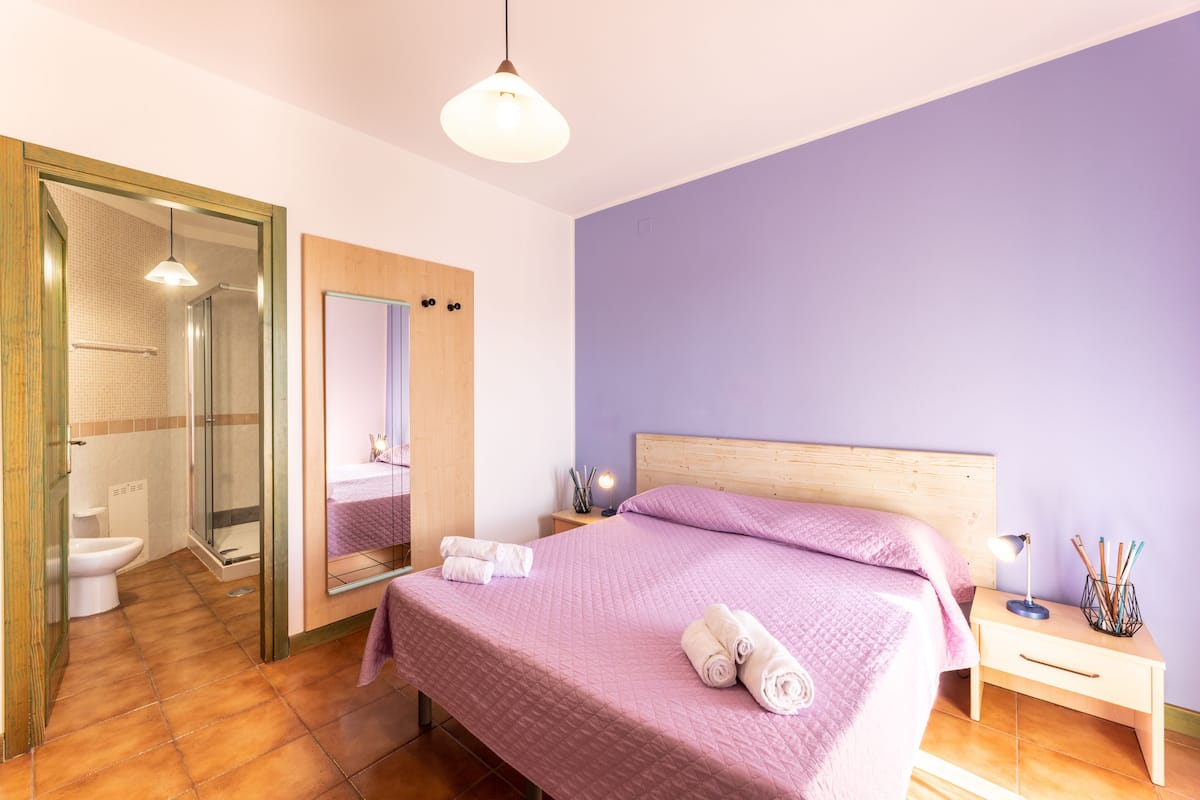A bedroom with a bed, a nightstand, a bedside lamp, and a bathroom with a toilet and shower.