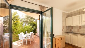 Una cocina moderna con armarios blancos y salpicadero de azulejos, un pequeño comedor con mesa y sillas, y un puerta corrediza de vidrio que da a un patio con mesa y sillas.