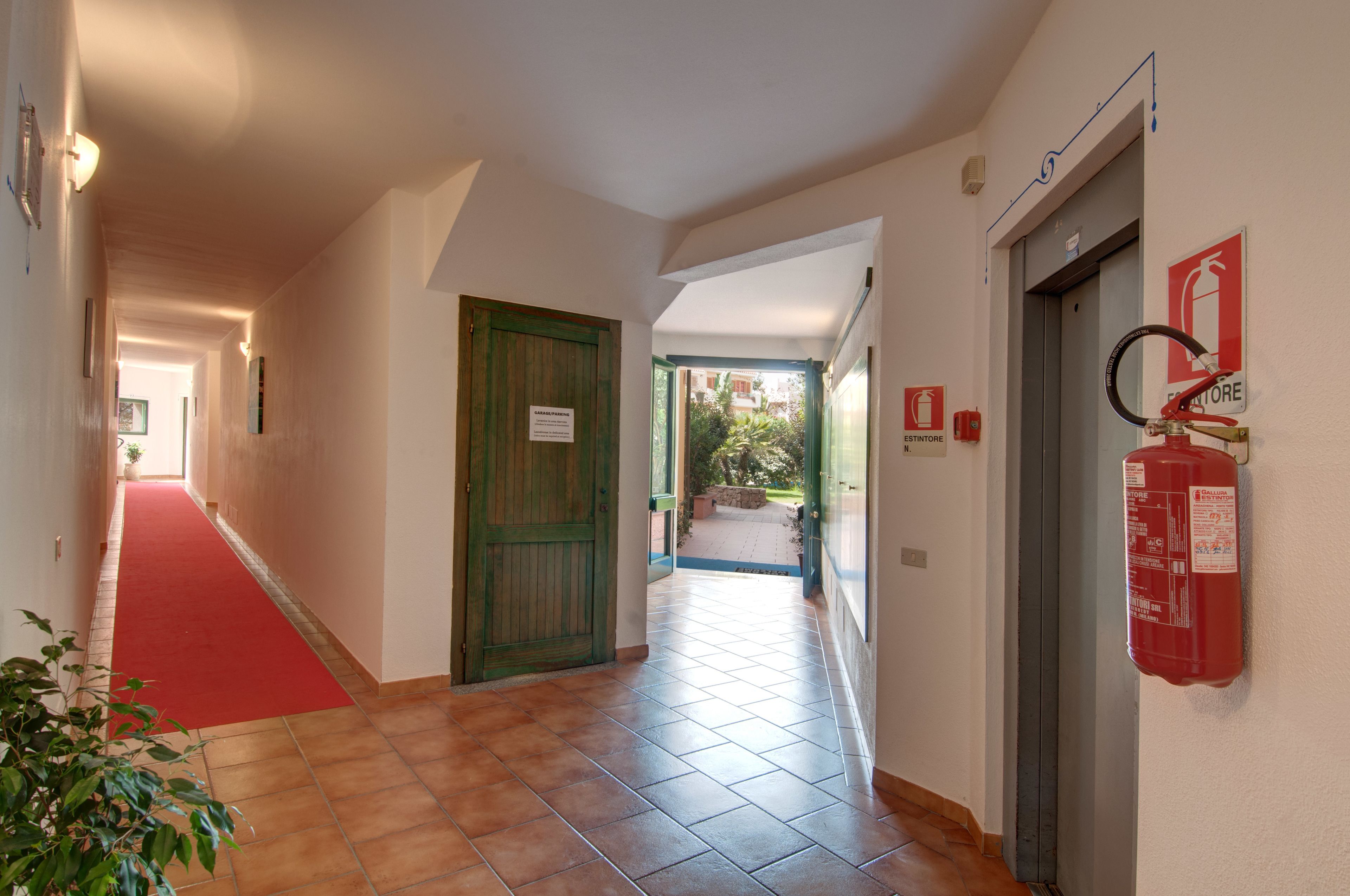A hotel corridor with a red carpet, tiled floor, green door, and a fire extinguisher mounted on the wall.