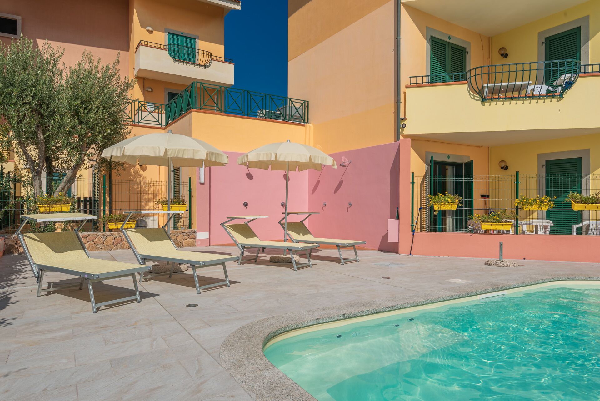 Área de piscina com espreguiçadeiras e guarda-sóis, piscina límpida e um edifício com varandas e plantas em vasos.