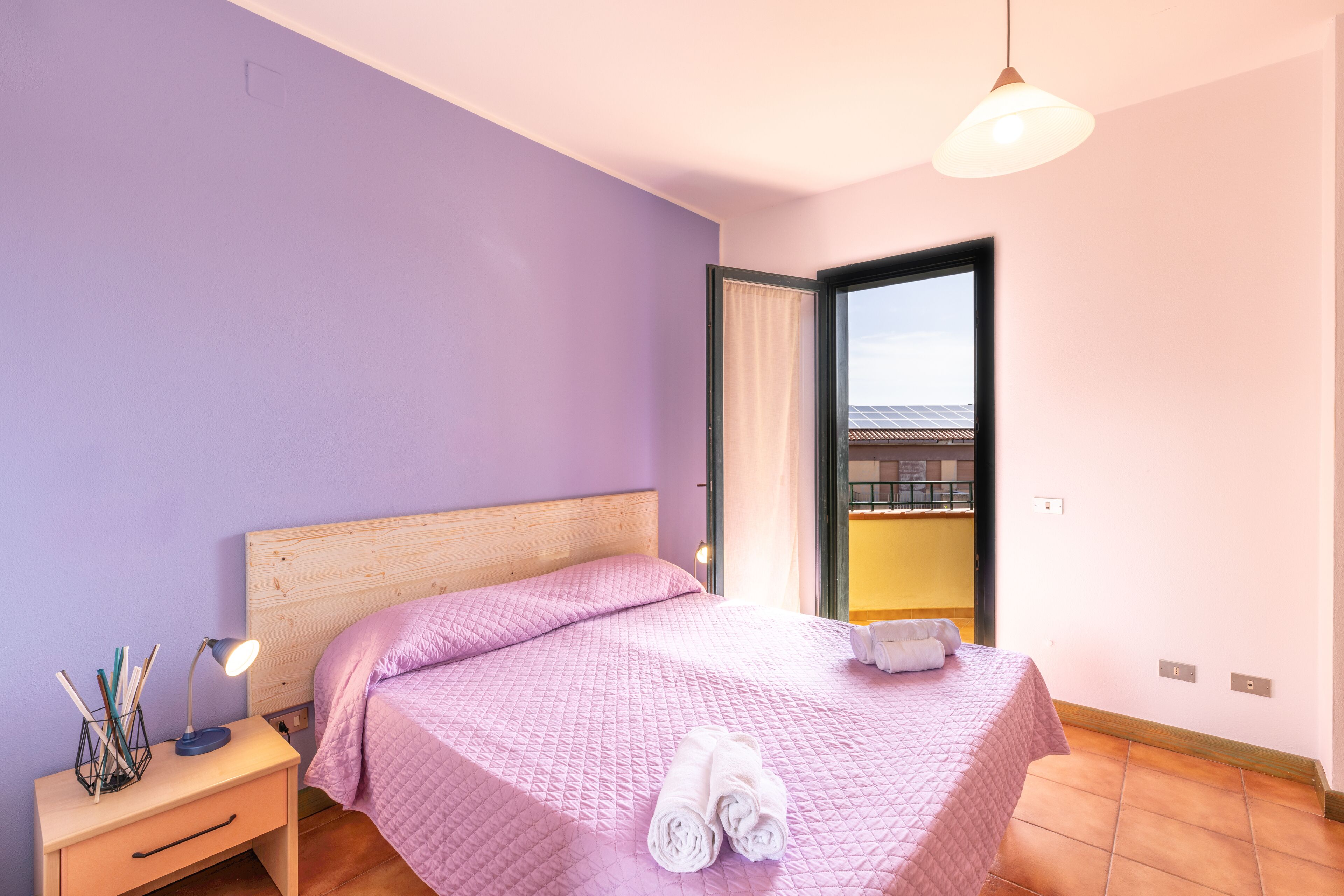 A bedroom with a wooden headboard, a pink quilted bedspread, a nightstand with a lamp, and a window with a view of a building.