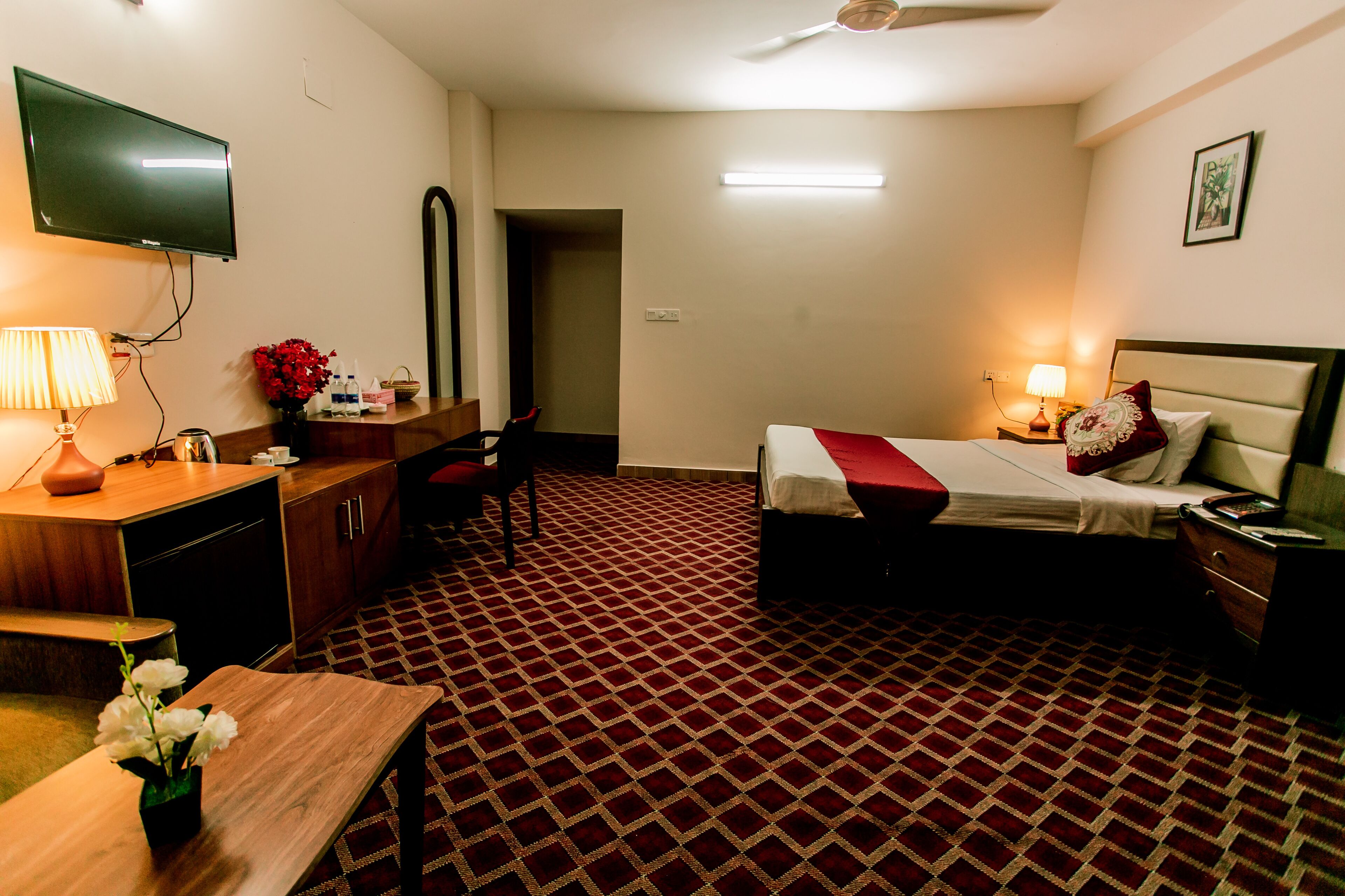 Ein Hotelzimmer mit Bett, Nachttisch, Lampe, Fernseher, Schreibtisch, Stuhl und einem gemusterten Teppich.