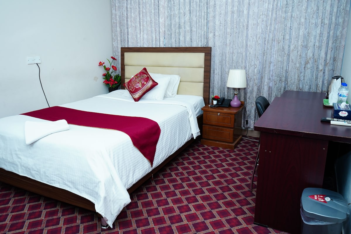 A hotel room with a bed, a desk, a chair, a waste bin, and a nightstand with a lamp.