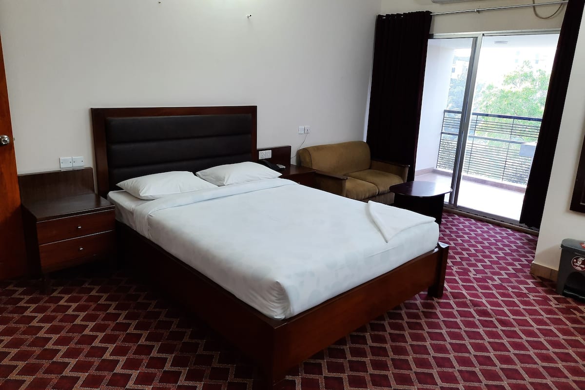 A hotel room with a bed, a nightstand, a chair, a small table, and a view of the outside through a sliding door.