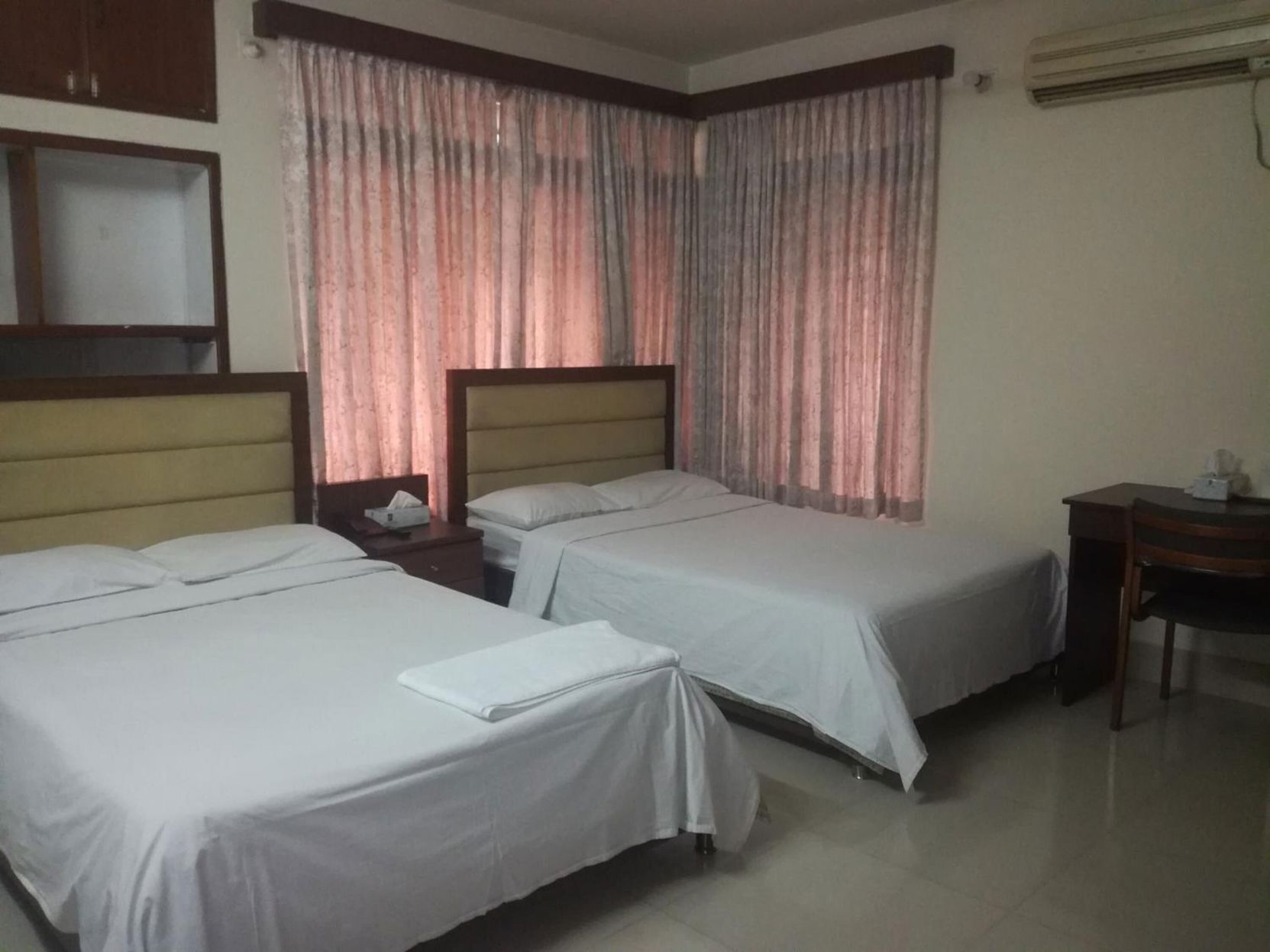 A hotel room with two single beds, a wooden headboard, a nightstand with a lamp, and a desk with a chair.