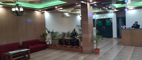 Un hall de hotel con piso de madera, sofás rojos, un mostrador de recepción y plantas en macetas.
