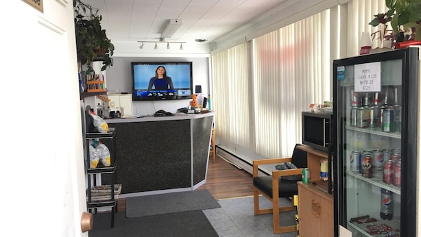 A reception area with a TV displaying a news broadcast, a counter, a refrigerator stocked with beverages, and a seating area with a chair and a small table. - Hilltop Motel (Kingston)