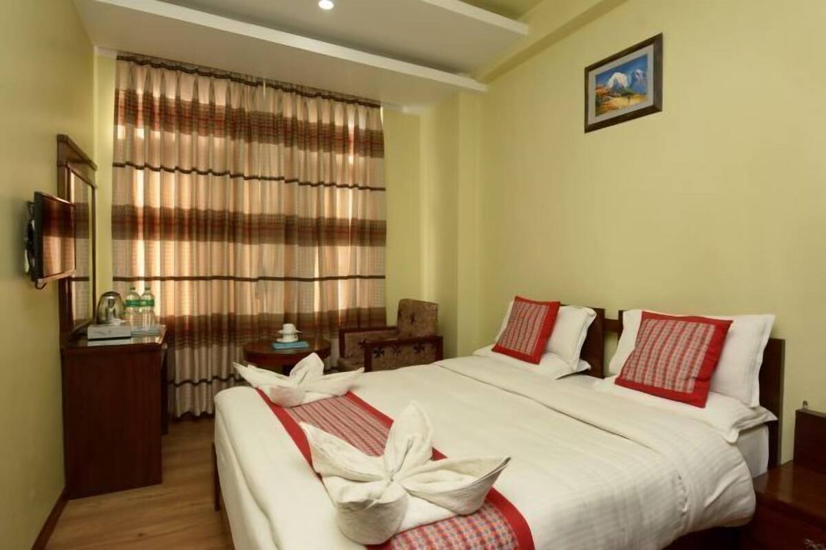 A hotel room with a bed, a desk with a chair, a small table with a lamp, and a painting on the wall.