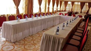 Una larga mesa de conferencias con botellas de agua y tarjetas de identificación, rodeada de sillas rojas en una sala con piso estampado y cortinas.
