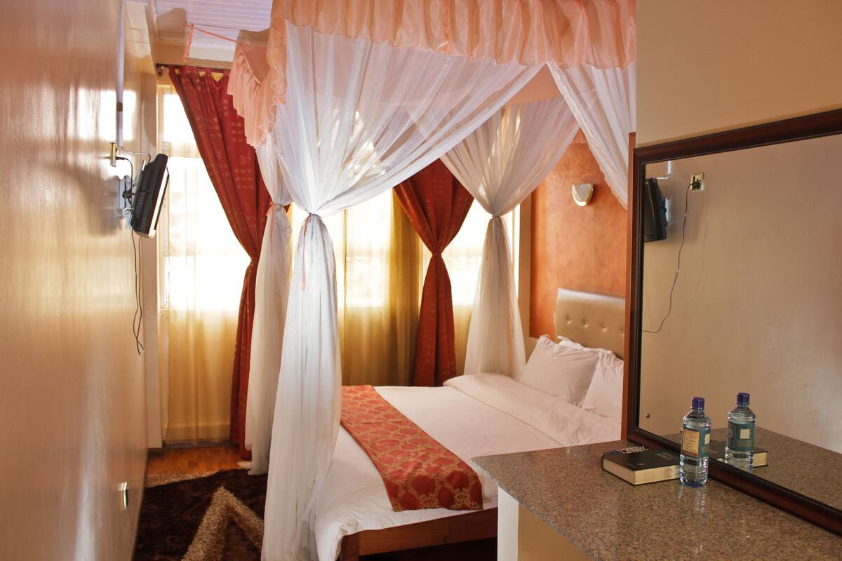 A hotel room with a bed, a bedside table, a mirror, and a window with curtains.