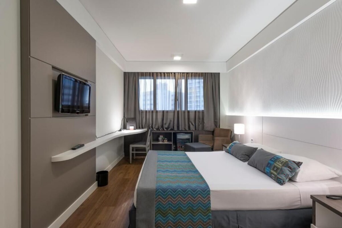 Et moderne hotelværelse med en seng, et fjernsyn ophængt på væggen, et skrivebord med stol og et vindue med gardiner.