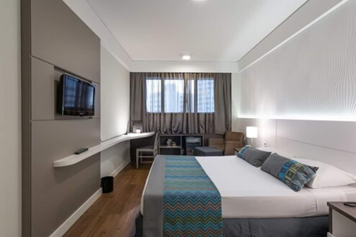 Ein modernes Hotelzimmer mit einem Bett, einem Schreibtisch, einem Fernseher und einem Fenster mit Vorhängen.