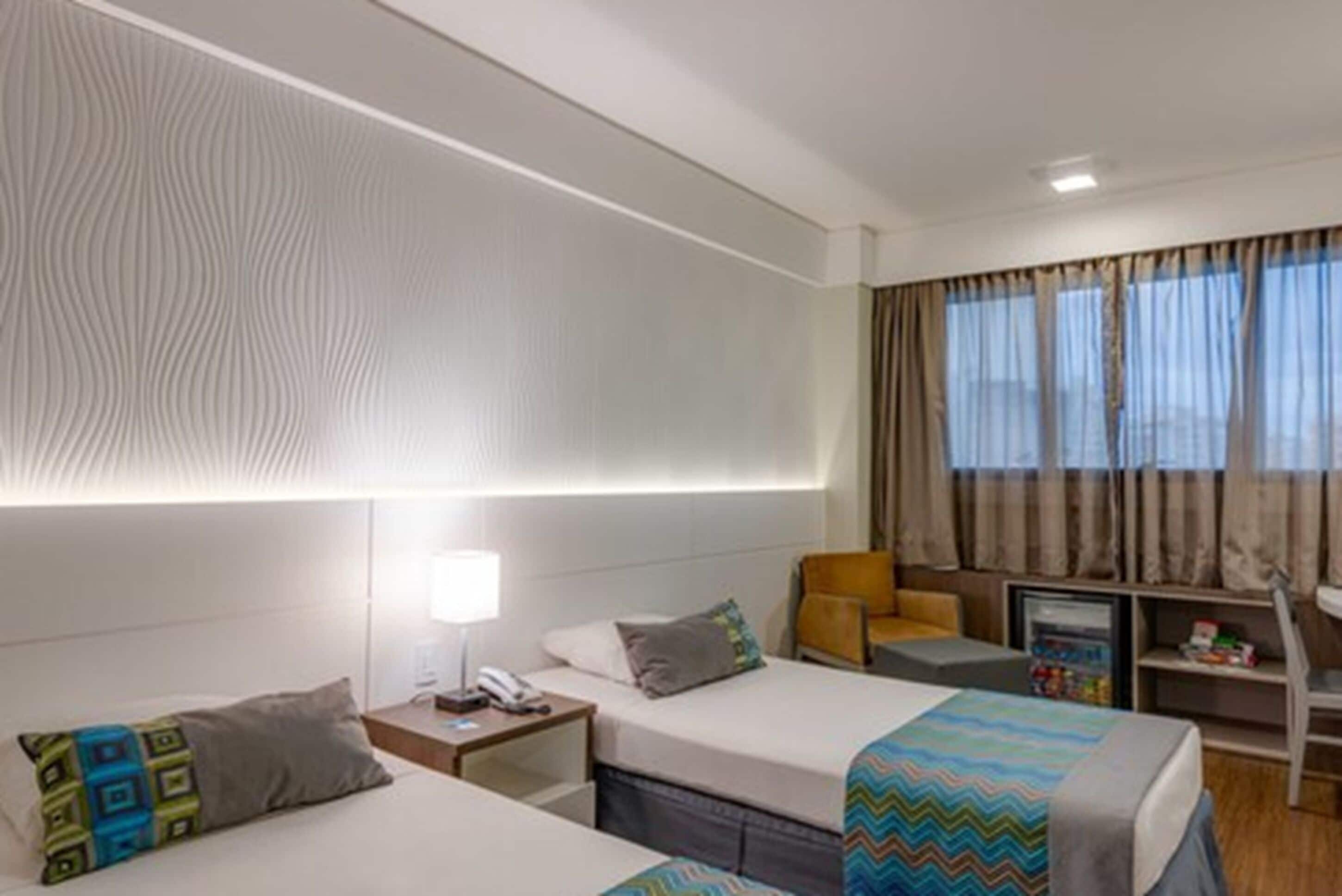 Ein Hotelzimmer mit zwei Betten, einem Schreibtisch, einem Sessel, einem Fernseher und einem Fenster mit Vorhängen.