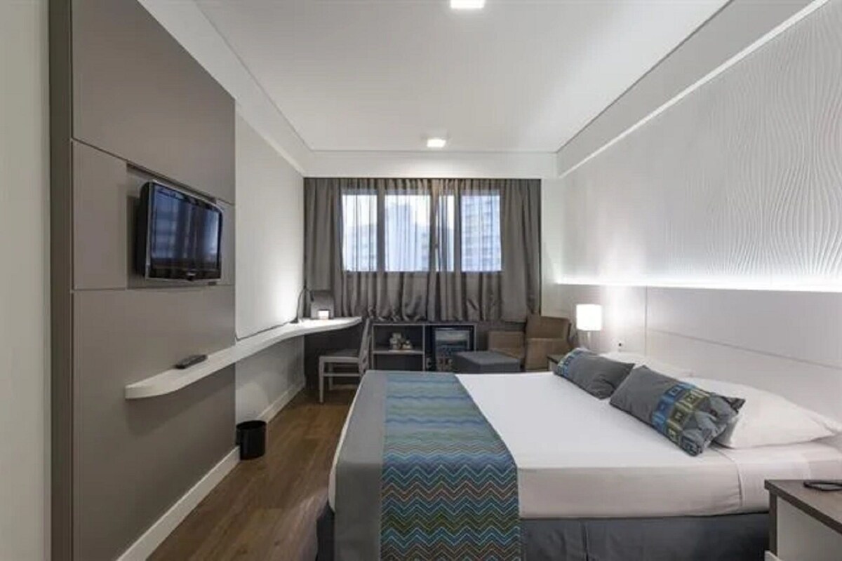 Et hotelværelse med seng, tv monteret på væggen, skrivebord med stol og et vindue med gardiner.