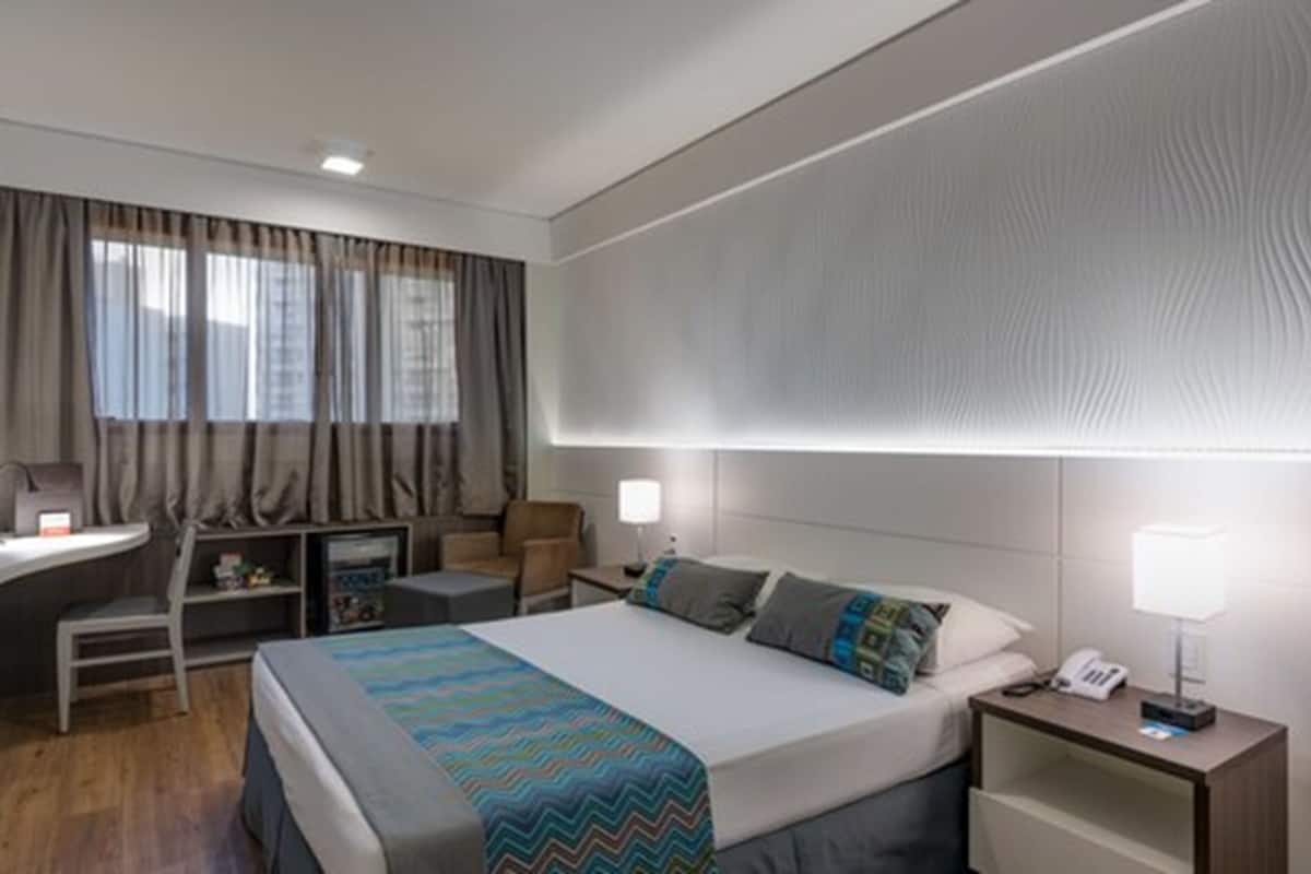 Ein Hotelzimmer mit Bett, Nachttisch, Lampe, Schreibtisch, Stuhl und Fernseher.