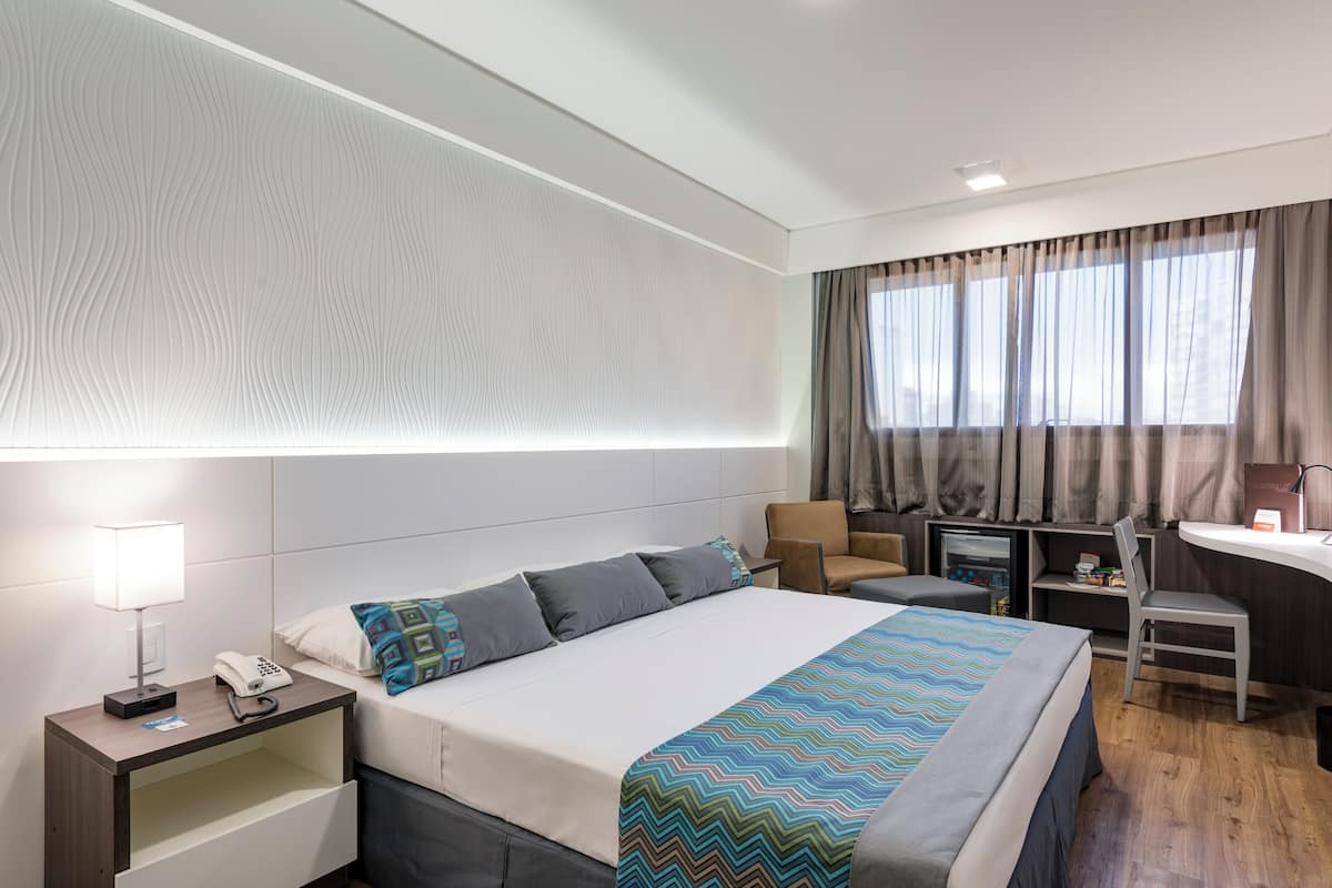 Ein Hotelzimmer mit einem großen Bett, einem Schreibtisch, einem Stuhl, einem Nachttisch mit Lampe und Telefon sowie einem Fenster mit Vorhängen.