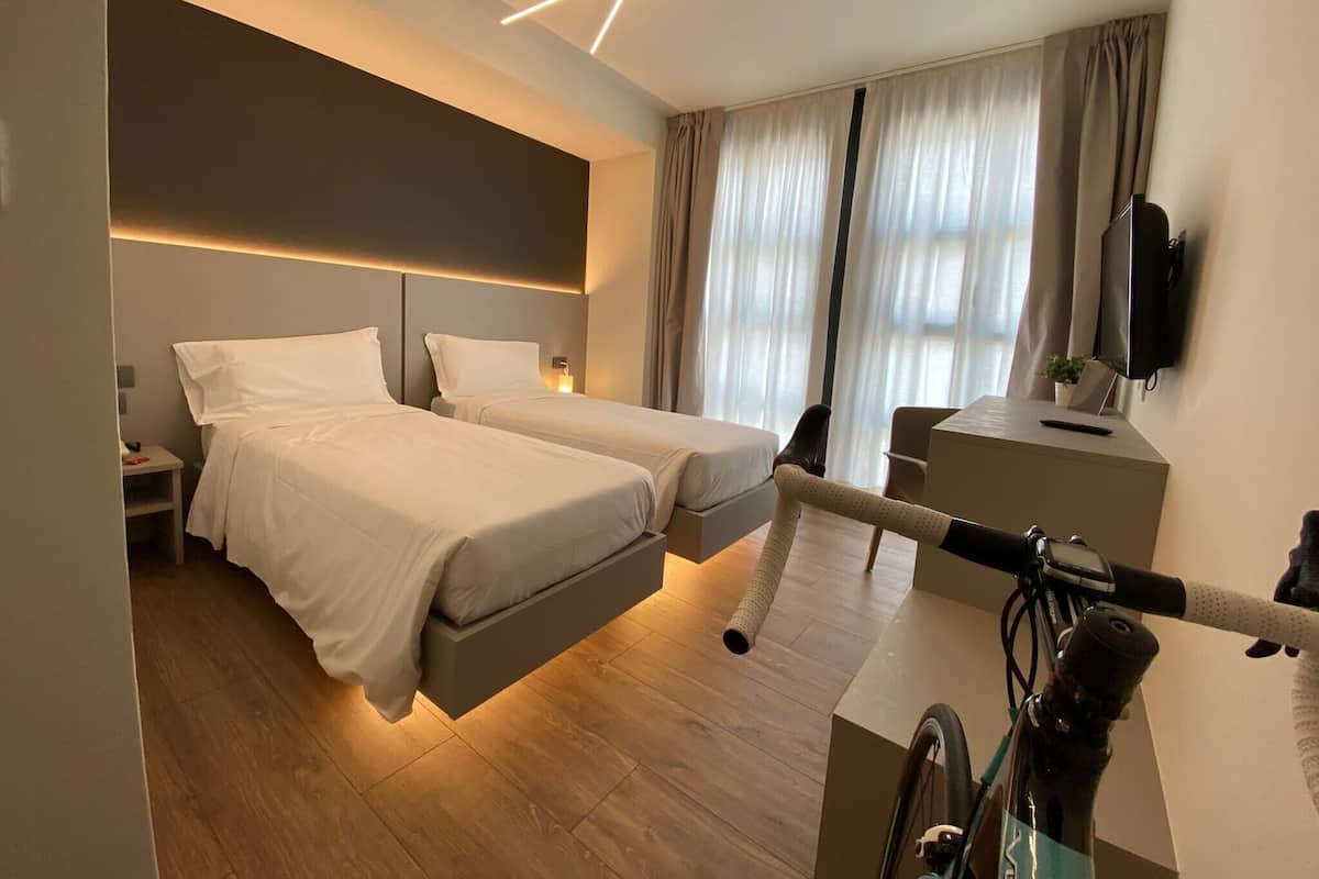A modern hotel room with two beds, a desk, a TV, and a bicycle.