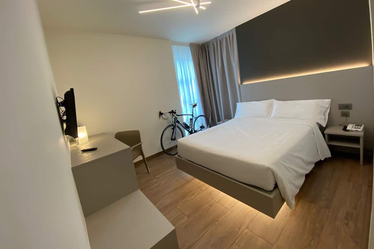 A modern hotel room with a bed, bedside table, lamp, bicycle, and a TV mounted on the wall.