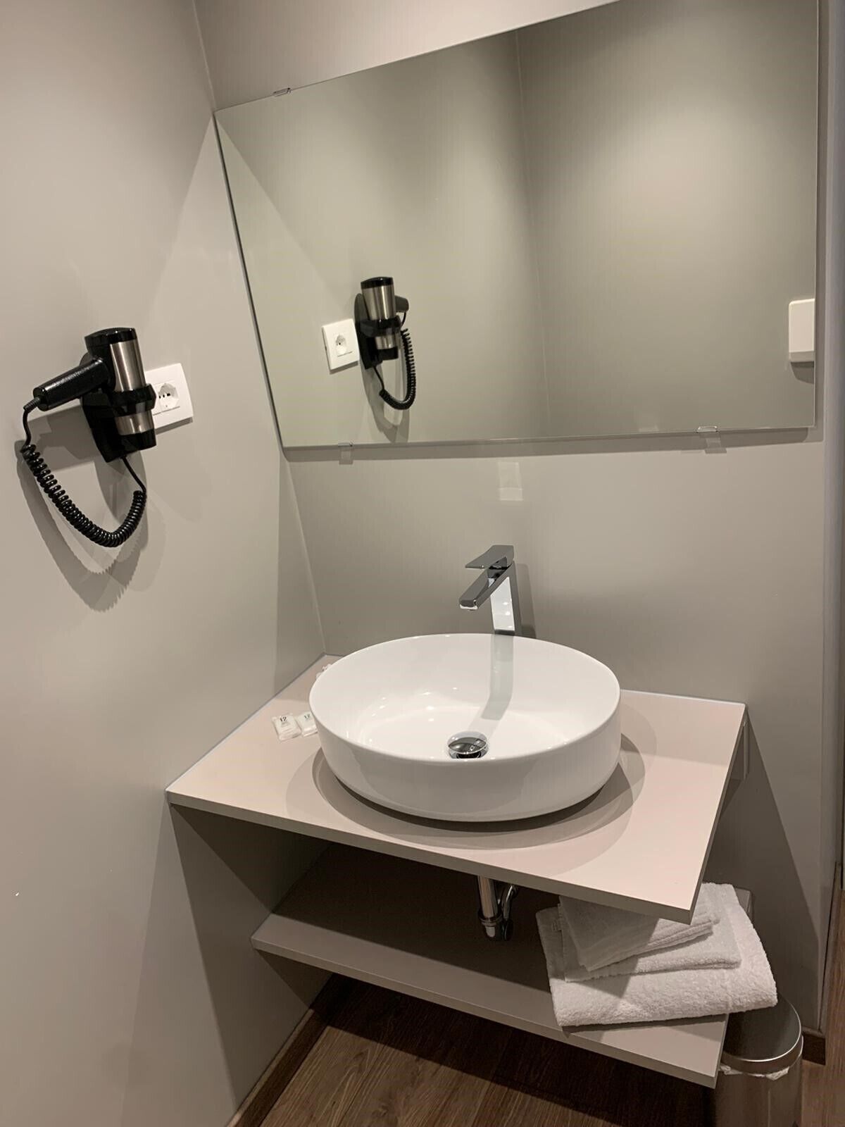 Ein modernes Badezimmer mit einem runden weißen Waschbecken, einem Spiegel darüber und einem an der Wand montierten Föhn.