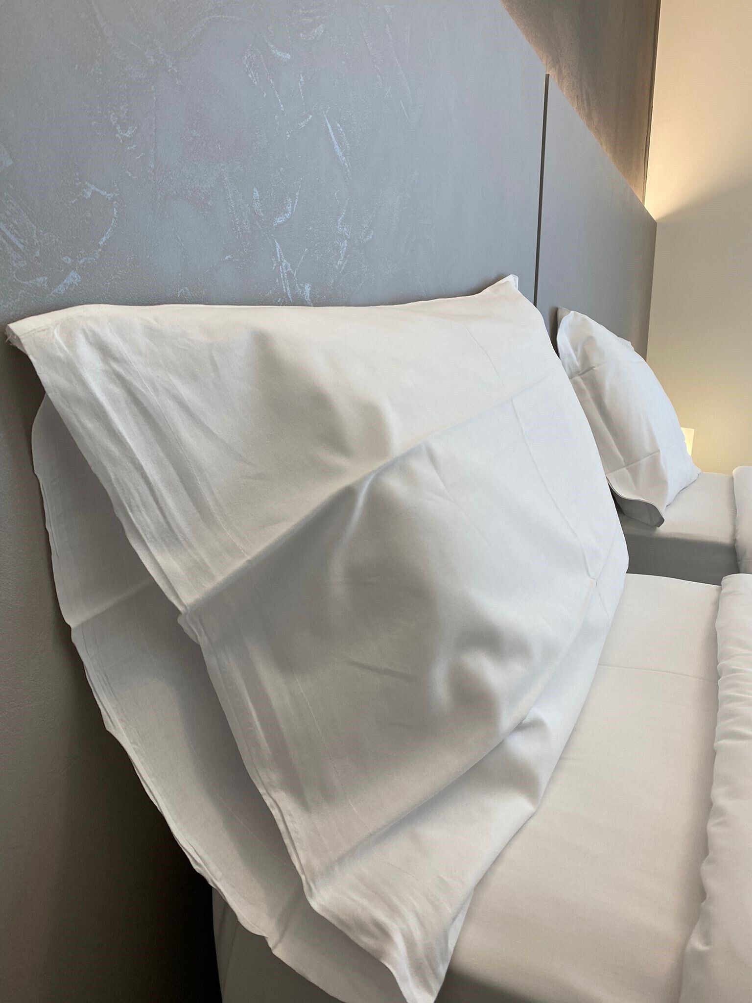 A neatly made bed with white linens and pillows, a grey textured wall, and a modern headboard.