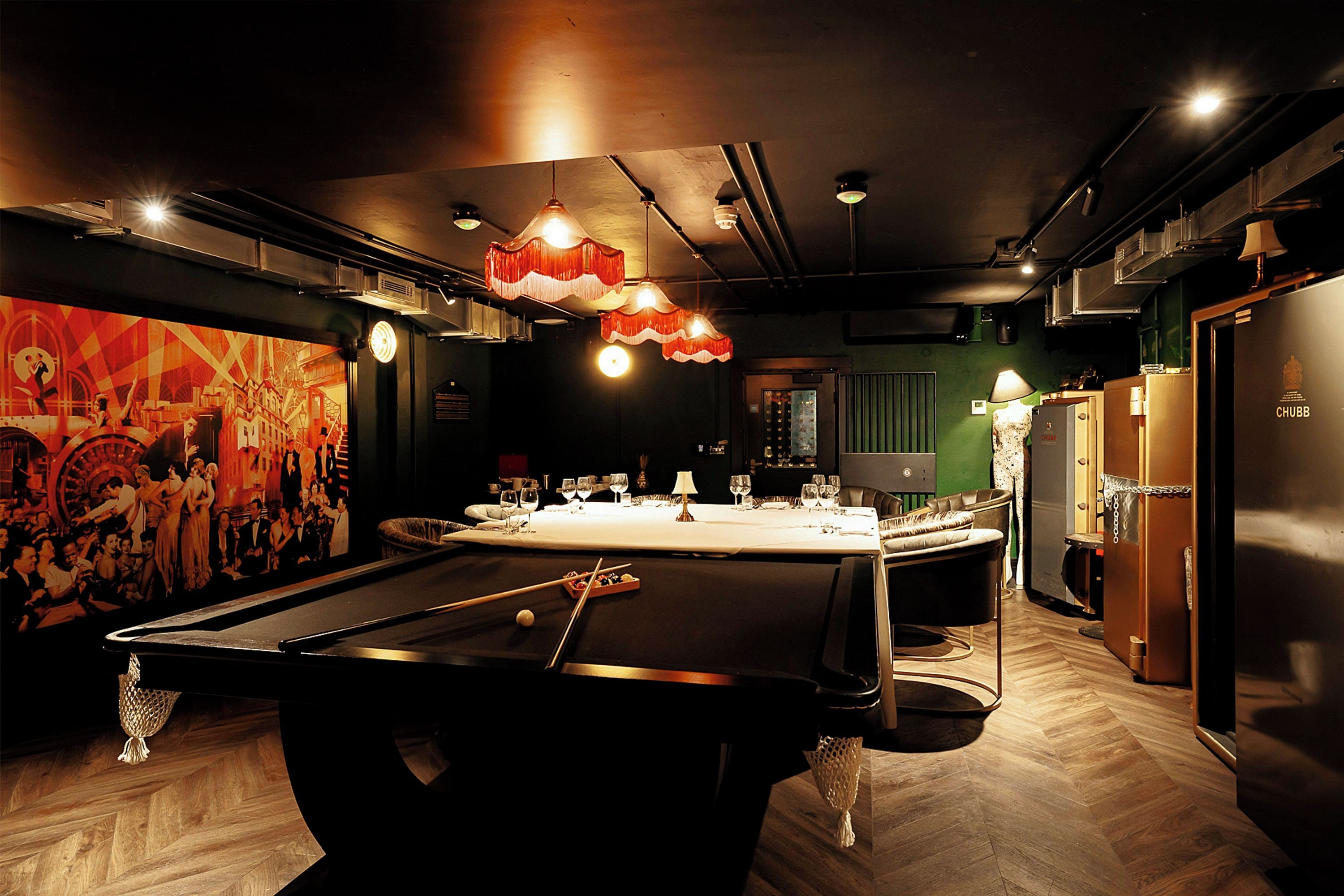 A billiards table with pool cues and balls, a bar area with stools, and a green wall with a mural.