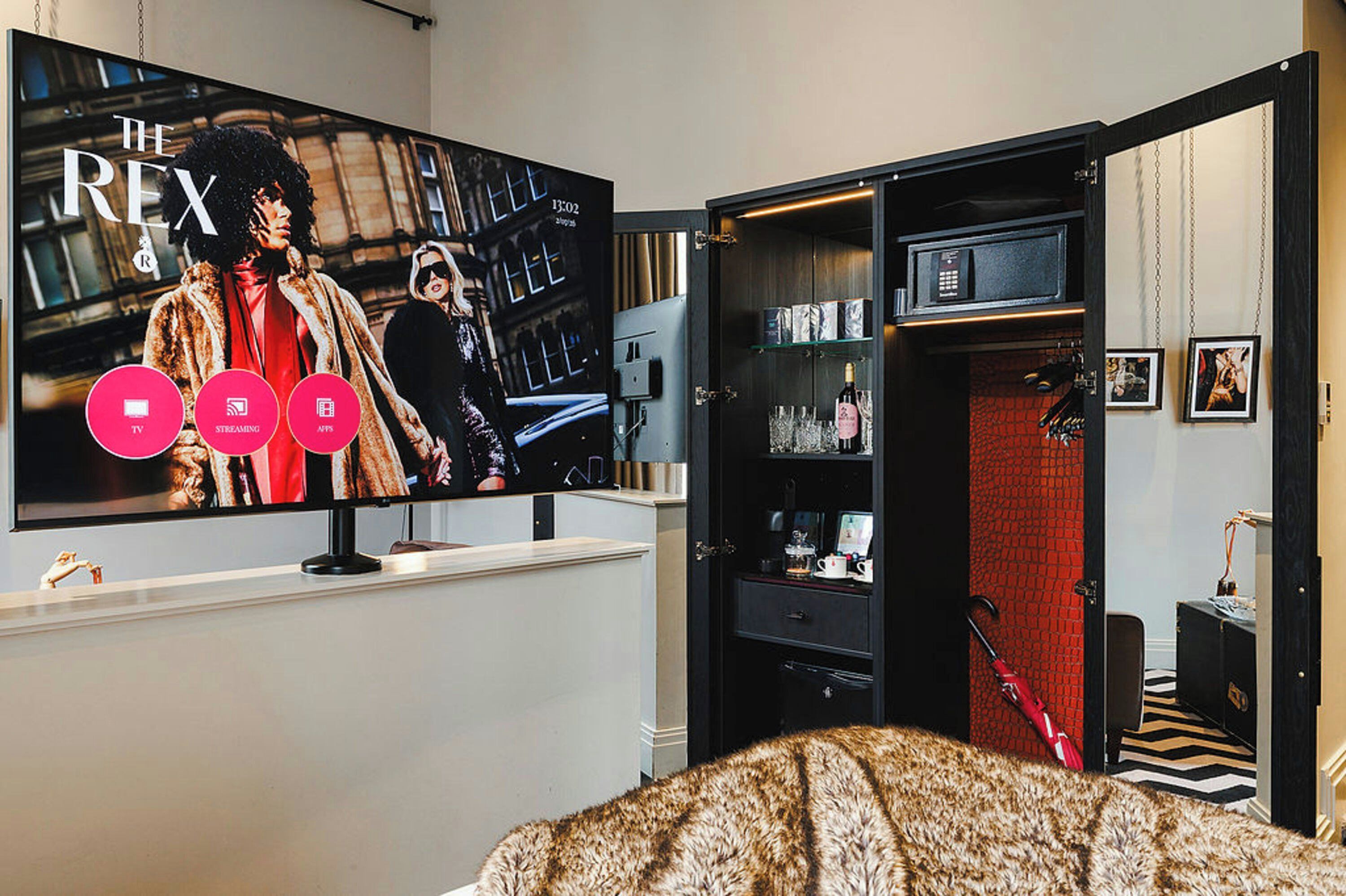 A room with a large flat-screen TV mounted on the wall displaying a movie poster. There is a black entertainment unit with shelves and a red carpeted area.