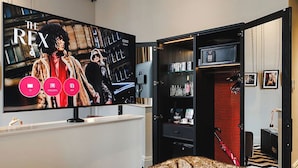 Habitación con un televisor de pantalla plana grande que muestra 'The Rex' e íconos para TV, streaming y aplicaciones. Se observa un armario con interior rojo, una cama con una colcha estampada y una pequeña mesa con una silla roja.