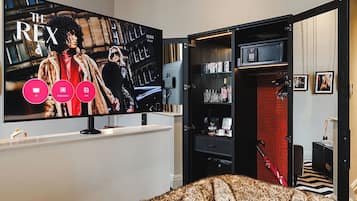 A room with a large flat-screen TV displaying 'The Rex' and icons for TV, streaming, and apps. There's a wardrobe with a red interior, a bed with a patterned blanket, and a small table with a red chair.