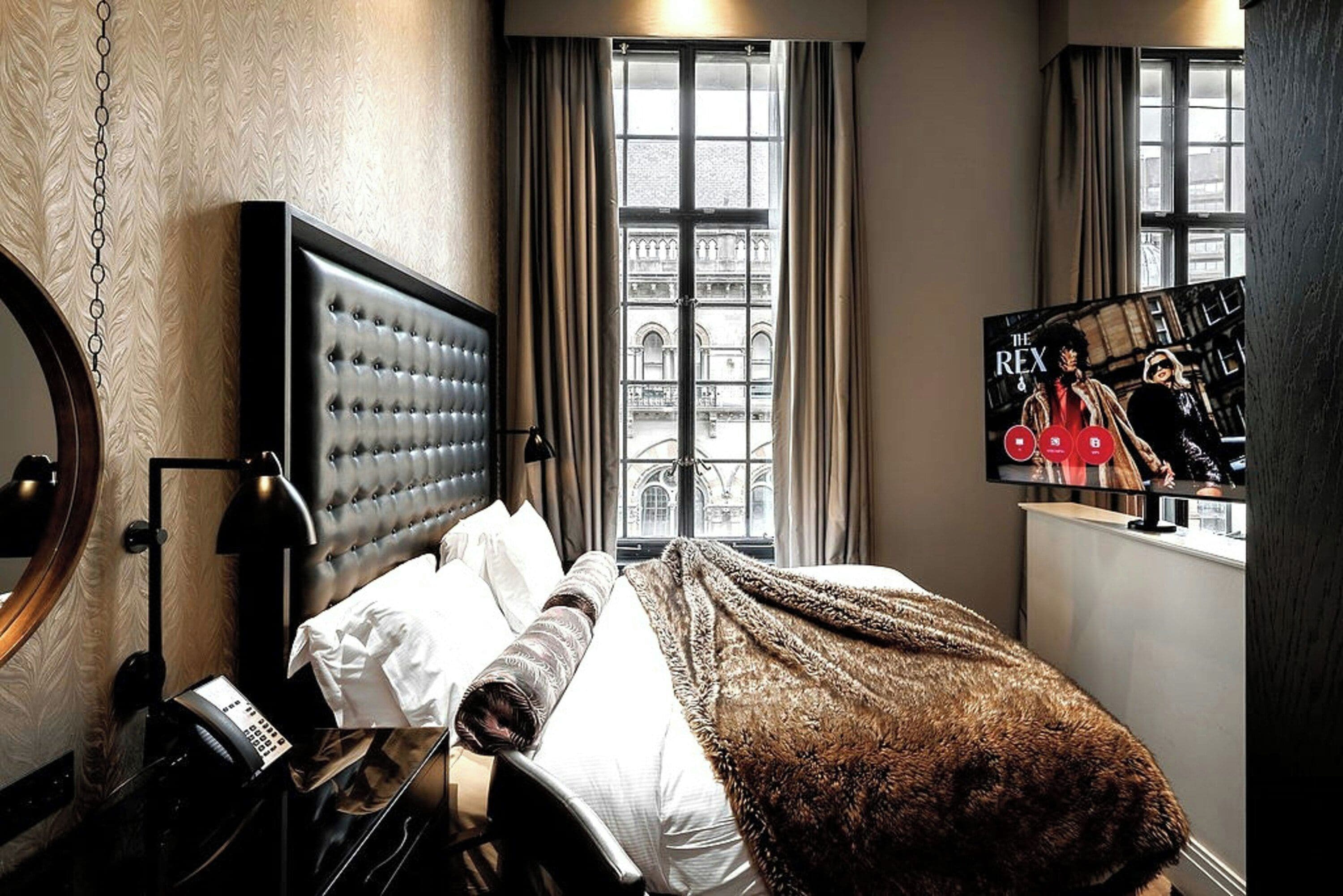 A hotel room with a large bed, a television, a window with a view of buildings, and a lamp.