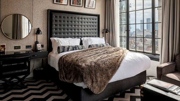 Habitación de hotel con una cama grande, vistas a la ciudad, un espejo redondo y un escritorio con una lámpara.