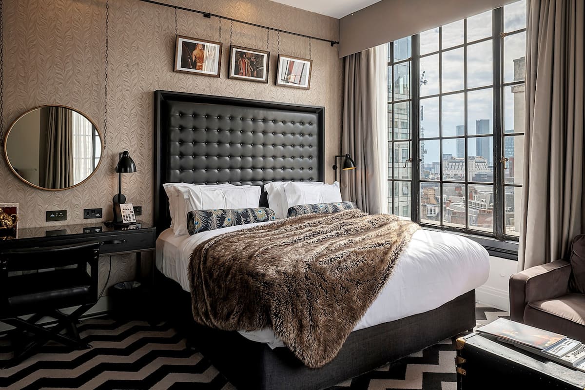 A hotel room with a large bed, a city view, a round mirror, and a desk with a lamp.