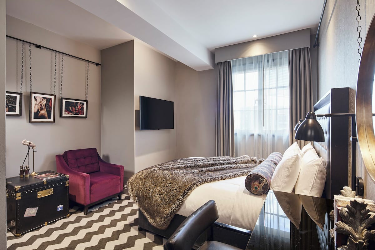 A hotel room with a bed, a desk, a chair, a dresser, a TV, and artwork on the wall.