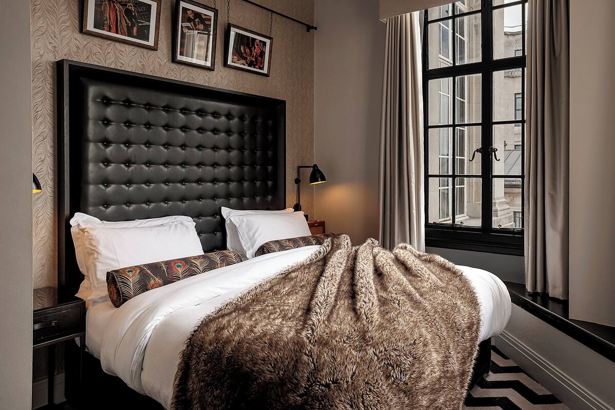 A bed with a tufted headboard, a patterned throw blanket, and a view of buildings through a window.