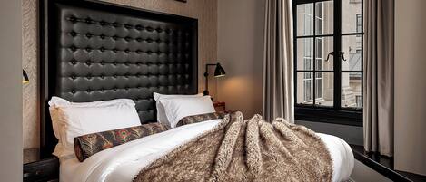Una cama con cabecera capitonada, una manta estampada y vistas de edificios a través de una ventana.