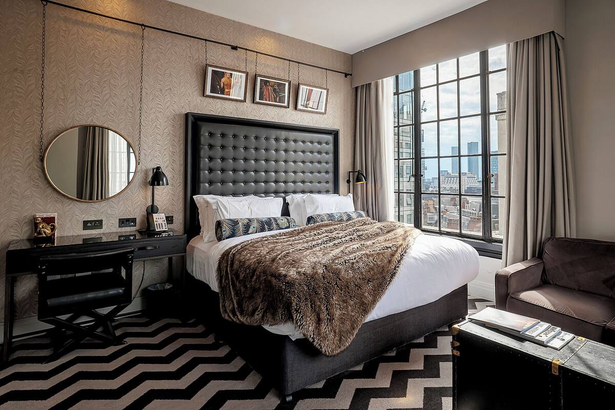 A hotel room with a large bed, a chair, a desk, a mirror, and a city view.