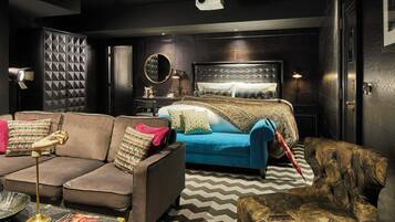 A modern living room with a sofa, a blue armchair, and a patterned armchair. There is a bed with a tufted headboard and a mirror above it.