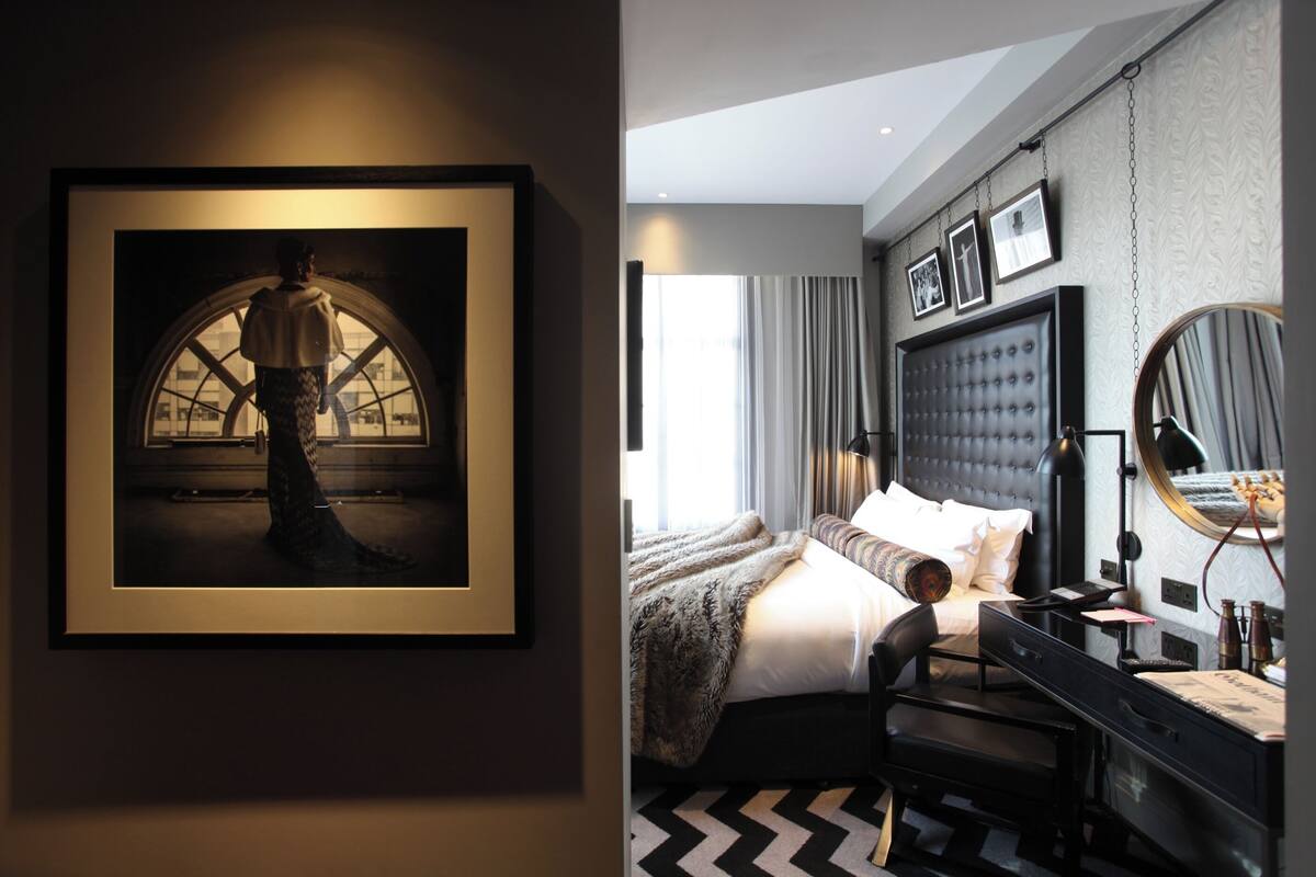 A modern hotel room with a large bed, a desk, a chair, a mirror, and a framed picture on the wall.