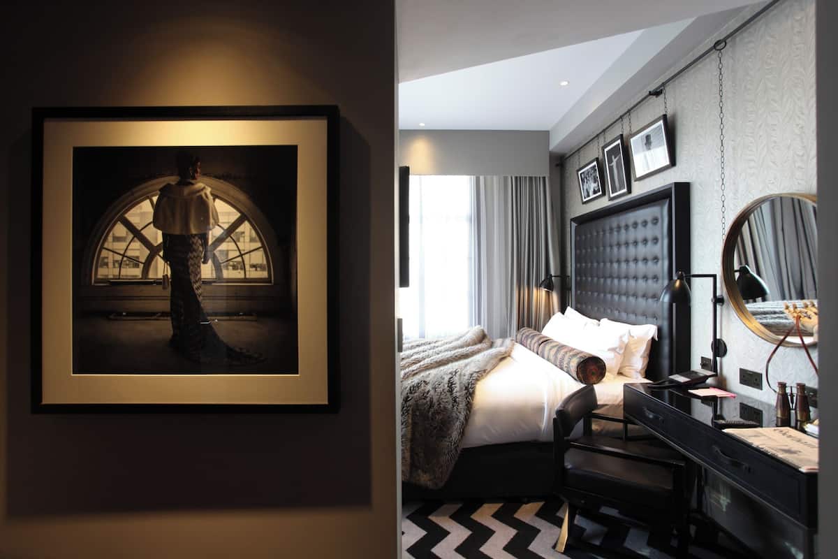 A modern hotel room with a large bed, a desk, a chair, a mirror, and a framed picture on the wall.