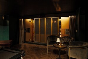 A dimly lit room with a wooden floor, a sofa, a chair, and a table with a lamp. - The Rex – Formerly Hotel Gotham (Manchester)