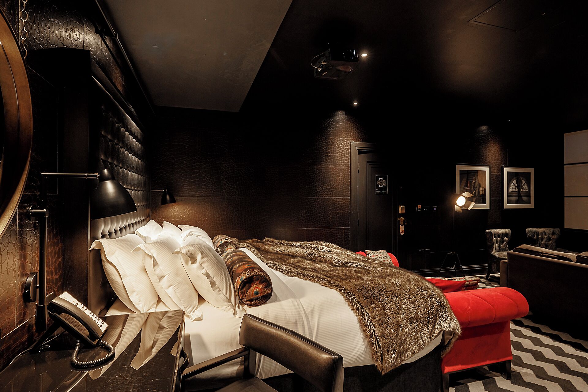 A hotel room with a bed, a red armchair, a telephone, and a wall-mounted safe.