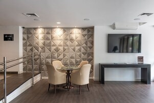 Una sala de conferencias moderna con pared de diseño geométrico, una mesa de vidrio con cuatro sillas, un aparador negro y un televisor de pantalla plana montado.