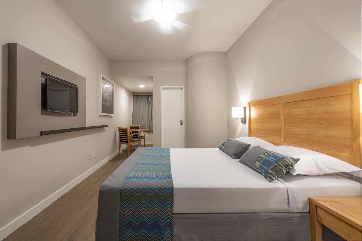 A hotel room with a bed, a TV mounted on the wall, a wooden headboard, and a small table.