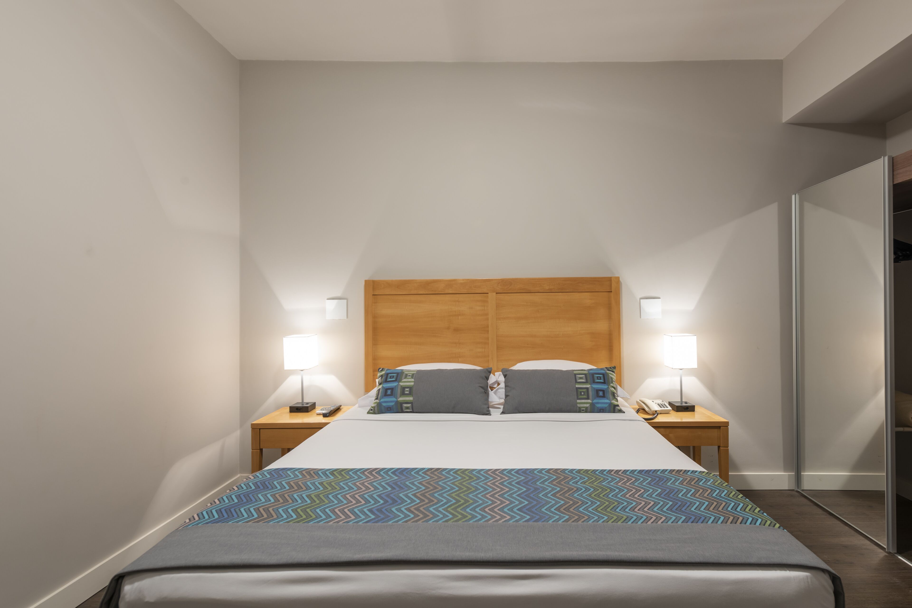 A hotel room with a large bed, two bedside tables with lamps, a wooden headboard, and a patterned bedspread.