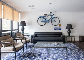 Una sala de estar moderna con un sofá de cuero negro, dos sillones, una mesa de centro de vidrio y un tapete azul estampado. Una bicicleta azul está colgada en la pared, y hay dos lámparas sobre mesas auxiliares.