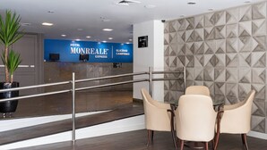 Zona de recepción de hotel con un letrero azul que dice "HOTEL MONREALE EXPRESS GLICÉRIO CAMPINAS", un mostrador de recepción, una planta en maceta y un área de descanso con tres sillas.