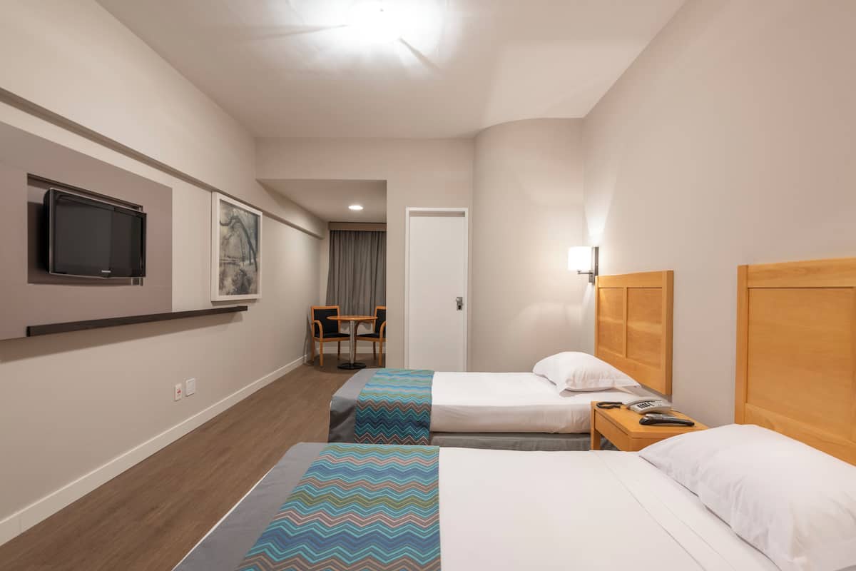 A hotel room with two beds, a TV mounted on the wall, a desk with a lamp, and a chair.