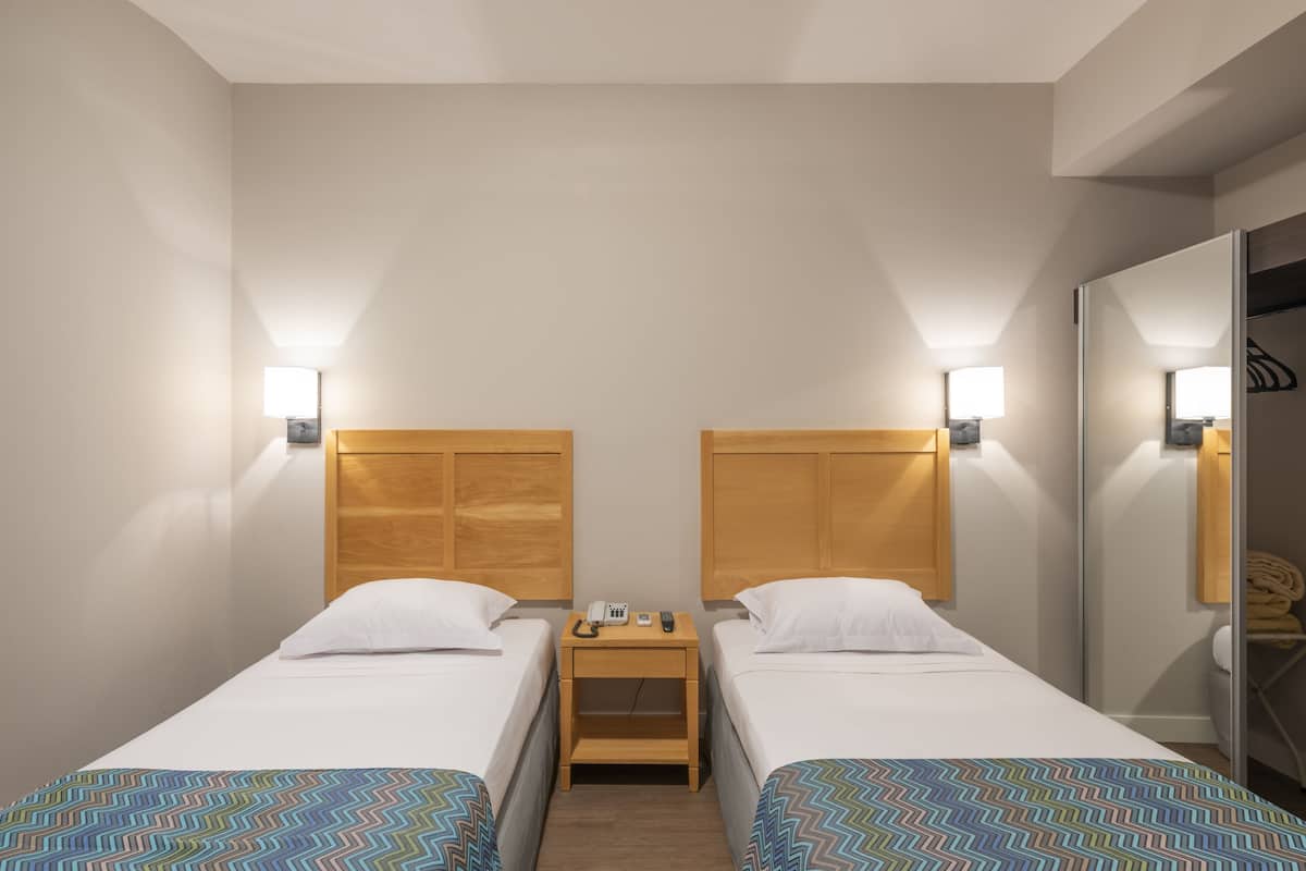 A hotel room with two single beds, a wooden nightstand with a phone and remote, and wall-mounted lights.