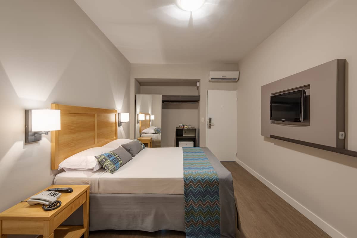 A hotel room with a bed, bedside table with a telephone, a TV mounted on the wall, and an air conditioning unit.